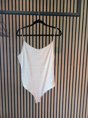 Vit ribbad body med smala axelband - Snygg vit body med smala axelband och ribbad struktur. Modellen har en enkel, stilren design med rundad urringning och tryckknappar i grenen. Perfekt att styla till jeans eller kjol för en clean look.