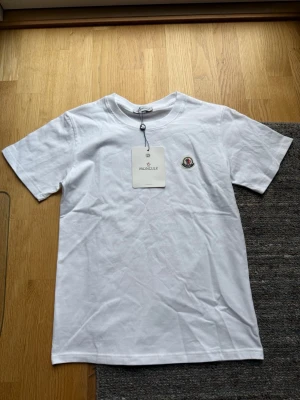 Vit Moncler t-shirt - 399kr! Köp båda för 649! Säljer en klassisk vit t-shirt från Moncler med rund hals och korta ärmar. T-shirten har Monclers ikoniska logga broderad på bröstet och är tillverkad i mjuk bomull för en skön känsla. Perfekt till jeans eller shorts för en clean look.
