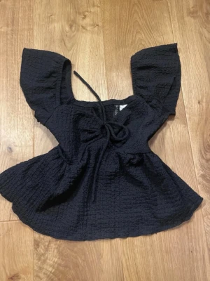 Svart topp med knyt och volangärm - Trendig svart topp från H&M med volangärmar och knyt framtill. Tillverkad i ett strukturerat bomullsliknande material. Toppen har en luftig passform och fyrkantig ringning som ger en modern look.