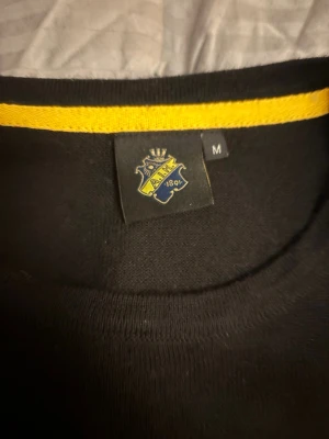 Svart tröja från aik  - Snygg svart tröja från aik  med rund halsringning och diskret logga på bröstet. Tröjan har en gul detalj på insidan av kragen och är perfekt för en stilren look. Långärmad och i mjukt material.knappt använd priset kan diskutera