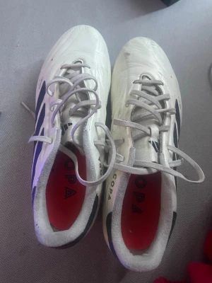 Vita Adidas Copa fotbollsskor - Säljer ett par vita Adidas Copa fotbollsskor med svarta detaljer och klassisk design. Skorna har snörning och röd innersula med Copa-tryck. Tillverkade i syntetmaterial för bra komfort och grepp på planen.