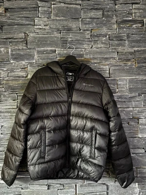 Svart pufferjacka från Champion - Säljer en svart pufferjacka från Champion med huva och dragkedja framtill. Jackan har två sidofickor och quiltad design med breda kanaler. Perfekt för kalla dagar och riktigt snygg till stilren-stil.