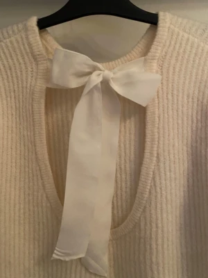 Krämvit stickad tröja med rosett - Supersöt krämvit stickad tröja med djup urringning i ryggen och en stor dekorativ rosett upptill. Tröjan har ribbstickad struktur och långa ärmar. Perfekt för dig som gillar en romantisk och trendig look. Superfint knyte i ryggen som ger en mer elegant intryck. 