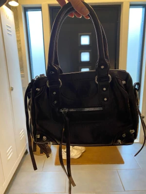 Mörkbrun handväska med nitar - Säljer en mörkbrun handväska i glansigt läder med silverfärgade nitar och detaljer. Väskan har dubbla handtag, dragkedja upptill och en liten ficka med dragkedja framtill. Dekorativa remmar hänger från dragkedjorna för en cool look.