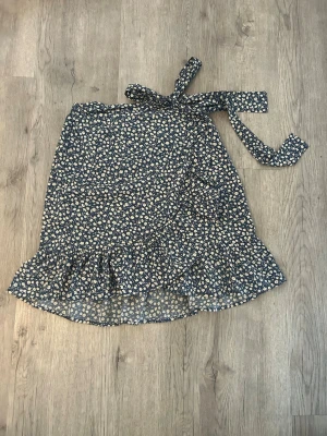 Blommig omlottkjol med volang, ONLY - Supersöt kort omlottkjol med blå botten och små vita blommor. Kjolen har volangkant nedtill och knytband i midjan för en justerbar passform. Perfekt för vår och sommar när du vill ha en lätt och luftig look.