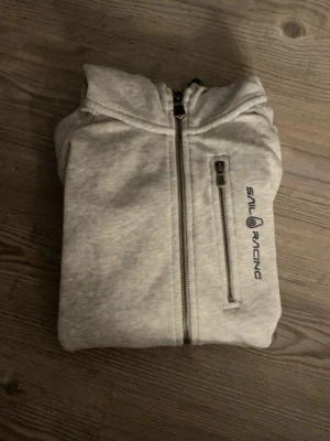 Ljusgrå hoodie från Sail Racing - Ljusgrå hoodie från Sail Racing med dragkedja framtill och en extra ficka med dragkedja på bröstet. Hoodien har en broderad logga på bröstet och huvan, samt mjukt material som känns skönt mot huden. 💓
