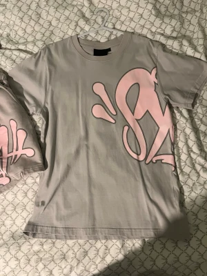 Syna world set  - Säljer ett par grå shorts med stort rosa graffiti-inspirerat tryck framtill. Shortsen har snörning i midjan med rosa dragsko och sidofickor. Perfekta för en avslappnad streetwear-look.
