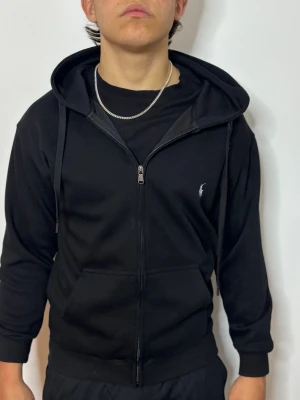 Svart zip hoodie från Polo Ralph Lauren - Säljer en svart hoodie med dragkedja från Polo Ralph Lauren. Klassisk design med huva, dragsnören och fickor framtill. Liten vit logga broderad på bröstet. Tillverkad i mjukt bomullsmaterial som är skönt att bära.