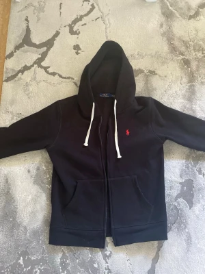 Svart hoodie från Polo Ralph Lauren - Svart hoodie med dragkedja från Polo Ralph Lauren. Klassisk design med vit dragsko i huvan och röd broderad logga på bröstet. Mjuk bomullskänsla, två fickor fram och ribbade muddar vid ärmar och nederkant. Perfekt för en chill och stilren look.
