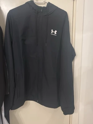 Svart vindjacka från Under Armour - Snygg svart vindjacka från Under Armour med huva och dragkedja framtill. Jackan har två sidofickor, justerbar dragsko i huvan och meshfoder för extra komfort. Under Armour-logga i vitt på bröstet ger en sportig look.
