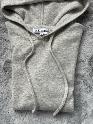 Ljusgrå hoodie från S&T Studios - Snygg ljusgrå hoodie från Studios i storlek S men den sitter som en XS. Tillverkad i mjukt material med ribbade muddar vid ärmslut och nederkant. Klassisk huva med dragsko och avslappnad passform, perfekt för en chill och stilren look. Lite smått defekt vid halsen men inget som märks