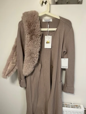 Beige kappa från Chiara Forthi - Stilren beige kappa från Chiara Forthi Milano med ribbad struktur och raka ärmar. Kappan har två fickor framtill och medföljer en mjuk, fluffig fuskpäls-krage i matchande ton. Perfekt för dig som vill ha en elegant och mysig look.