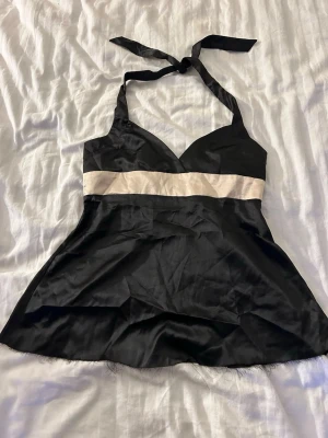 Svart satin nattlinne med halterneck - Snyggt svart nattlinne i satin med halterneck och bred vit rand under bysten. Mjuk och glansig känsla, lätt utställd passform och kort längd. Perfekt för en avslappnad kväll hemma eller som festtopp.