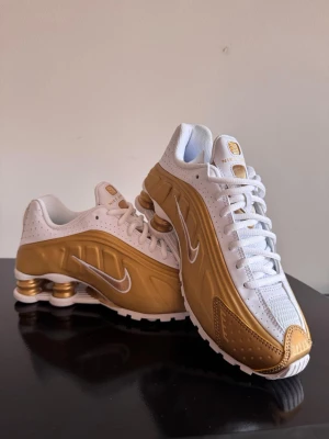 Nike Shox vita och guldiga sneakers - Nike Shox sneakers med vit mesh och läder, detaljer i metallic guld och vita snören. Ikoniska Shox-dämpning i hälen med guldiga fjädrar. Snygg design med perforeringar och silverfärgad Swoosh på sidan. Perfekt för dig som vill sticka ut med stil.