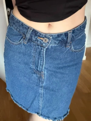 Blå jeanskjol  - Säljer en klassisk blå jeanskjol med rå, fransig kant nertill och femficksmodell. Kjolen har normal passform och stängs med knapp och dragkedja framtill. Perfekt till en avslappnad och trendig look.