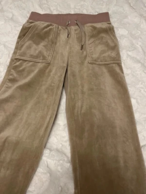 Beige velourbyxor från Juicy Couture - Säljer ett par beige mjukisbyxor i velour från Juicy Couture. Byxorna har bred resårmidja med dragsko, stora fickor fram och bakfickor med knapp. JC-logga broderad på ena fickan. Perfekta för en chill och trendig look.