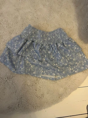 Blå blommig volangkjol - Söt blå kortkjol med vita små blommor och bred resår i midjan. Kjolen har dubbla volanger som ger en lekfull look och passar perfekt till sommaren. Materialet känns lätt och luftigt.