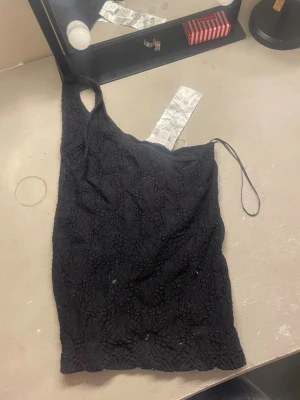 Svart oneshoulder topp med spetsdetaljer - Säljer en svart oneshoulder topp med snygga spetsdetaljer och strukturmönster. Toppen har en axelbandslös design på ena sidan och ett smalt axelband på andra. Perfekt för dig som vill sticka ut lite extra.