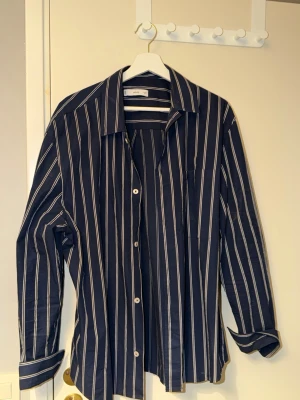 Mörkblå randig skjorta från MING - Snygg mörkblå skjorta från MING med vita vertikala ränder. Klassisk krage, långa ärmar och knäppning framtill. Tillverkad i mjuk bomull som känns skön mot huden. Perfekt för en clean och stilren look.