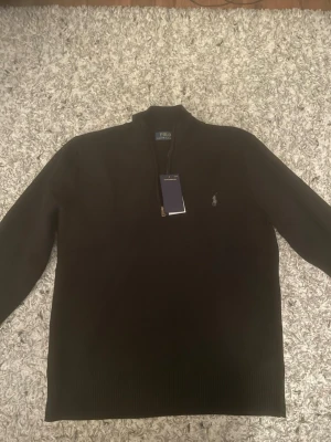 Svart stickad tröja från Polo Ralph Lauren - Säljer en svart stickad tröja från Polo Ralph Lauren med halvzip och klassisk logga broderad på bröstet. Tröjan har hög krage och ribbstickade muddar. Perfekt för dig som gillar stilrena och tidlösa plagg.
