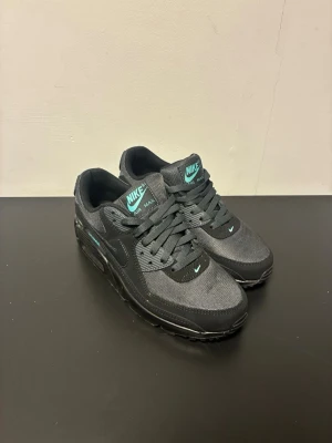 Nike Air Max 90 svart/turkos sneakers - Nike Air Max 90 sneakers i svart mesh och syntet med turkosa detaljer på plös, häl och sidor. Klassisk låg siluett med synlig Air-enhet i sulan och ikonisk Swoosh på sidan. Sköna och stilrena skor som passar perfekt till streetwear.