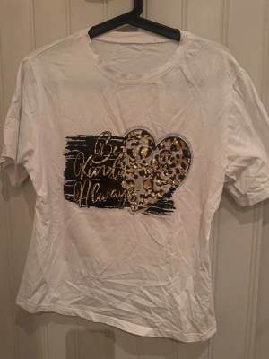 Vit t-shirt med leopardhjärta och paljetter - Snygg vit t-shirt med korta ärmar och ett stort hjärta i leopardmönster framtill. Texten 'Be Kind Always' i glittriga paljetter ger extra bling. Perfekt för dig som gillar djurmönster och detaljer som sticker ut.