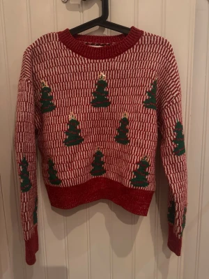 Röd jultröja med julgransmotiv - Stickad röd och vit tröja med gröna julgranar och guldfärgade stjärnor. Tröjan har rund halsringning och långa ärmar. Perfekt för julens alla tillfällen och har ett klassiskt julmönster som sticker ut.