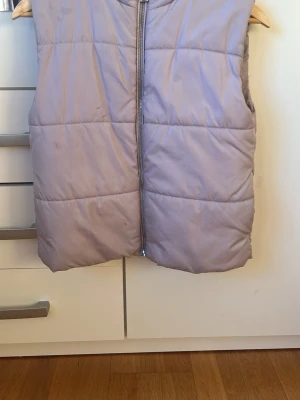 Ljuslila dunväst med dragkedja - Ljuslila dunväst med quiltad design och rak passform. Västen har en hel dragkedja framtill och är tillverkad i ett glansigt material. Perfekt för att hålla värmen under kyliga dagar. Reflekterande detalj på ryggen.