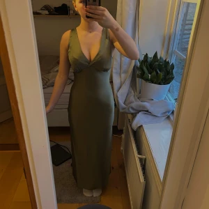 Grön långklänning med v-ringning - Snygg grön långklänning med djup v-ringning och breda axelband. Klänningen har en figurnära passform och är ärmlös, vilket ger en elegant siluett. Perfekt för dig som vill sticka ut med en stilren look. Materialet känns mjukt och följsamt mot kroppen.