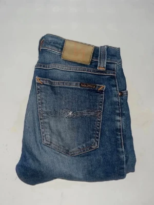 Nudie jeans  - Säljer ett par Nudie Jeans Tube Kelly i klassisk blå tvätt. Jeansen har raka ben och snygga kontrastsömmar. Tillverkade i ekologisk bomull med lite stretch för extra komfort. Midja 37cm, längd 108cm. JAG SKICKAR EJ FLER BILDER NÄR JAG BÄR JEANSEN! för storleksguide hänvisar jag till måtten och för bättre inblick i passformen rekommenderar jag att googla på modellnamnet. kolla måtten noggrant :) s5 