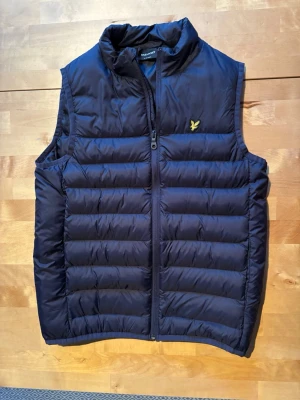 Mörkblå väst från Lyle & Scott - Mörkblå väst  från Lyle & Scott med quiltad design och hög krage. Västen har en gul logga på bröstet och dragkedja framtill. Två framfickor. Använd max tre gånger. Perfekt för lager på lager och ger en sportig look.