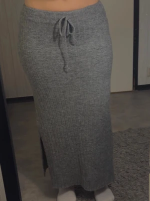 Grå ribbad stickad maxikjol - Mysig grå stickad maxikjol med ribbad struktur och dragsko i midjan. Kjolen har en slits på sidan som ger extra rörelsefrihet och en avslappnad look. Perfekt för dig som gillar bekväma och trendiga plagg till vardags.