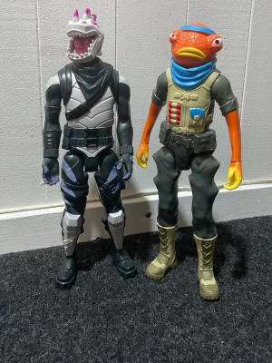 Fortnite actionfigurer, 2-pack - Två stora Fortnite actionfigurer i plast. Ena figuren har en hajmask med svart och vit dräkt, den andra har ett orange fiskhuvud, blå scarf och militärinspirerade kläder. Perfekta för samlare eller som dekoration i gamingrummet.