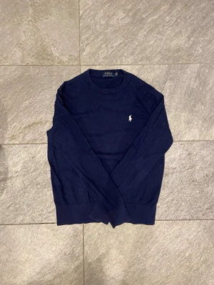 Mörkblå Kashmirtröja Polo Ralph Lauren - Mörkblå stickad tröja från Polo Ralph Lauren med rund halsringning och den klassiska broderade loggan i vitt på bröstet. Tröjan har långa ärmar och ribbade muddar vid ärmslut och nederkant. Perfekt för lager på lager och stilren look. 15% kashmir.