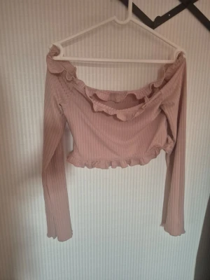 Rosa croppad offshoulder topp - Supersöt ljusrosa croppad topp med offshoulder-design och volangdetaljer längs halsringning och nederkant. Toppen är ribbad och har långa ärmar, perfekt för en trendig och feminin look.