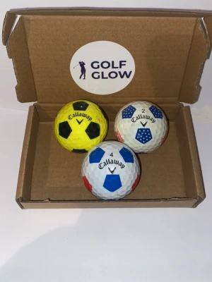Callaway chromesoft truvis 3-pack - Tre golfbollar från Callaway i en brun presentförpackning. En boll är gul med svarta fält, en vit med blå och röda fält, och en vit med blå stjärnor och röda detaljer. Perfekt för dig som vill ha unika golfbollar med cool design.