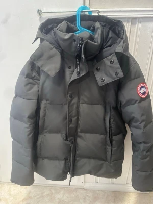 Canada Goose Wyndham - Snygg mörkgrå dunjacka från Canada Goose med klassisk patch på ärmen. Jackan har stor huva, hög krage och dragkedja med tryckknappar. Fylld med dun för extra värme och har två stora fickor framtill. Perfekt för kalla vinterdagar.