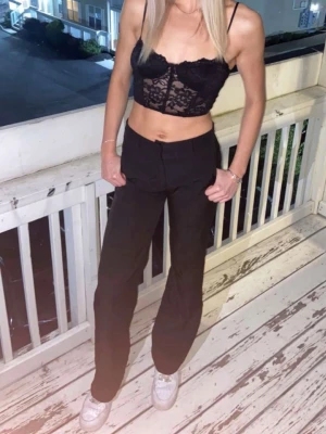 Svart spetsig croppad linnetopp - Svart croppad linnetopp med tunna axelband och spetsdetaljer. Toppen har en figurnära passform och är perfekt för en trendig look. Snyggt genombruten spets framtill och smala band ger en feminin känsla.