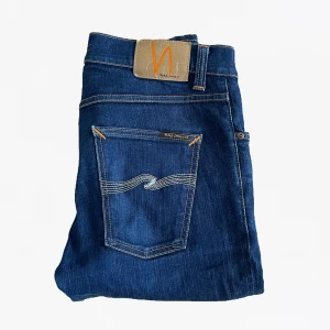 Mörkblå jeans från Nudie Jeans - Säljer ett par mörkblå jeans från Nudie Jeans med klassisk femficksmodell och kontrastsömmar. Jeansen har en rak passform och är tillverkade i denim med snygga detaljer på bakfickan och läderpatch i midjan.