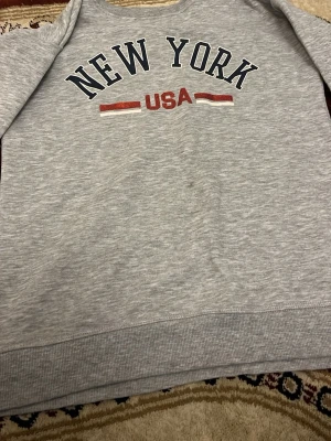 Grå sweatshirt New York USA - Snygg grå sweatshirt med trycket 'NEW YORK USA' i svart och rött framtill. Tröjan har rund halsringning och ribbade muddar vid ärmslut och nederkant. Perfekt för en avslappnad streetwear-look.