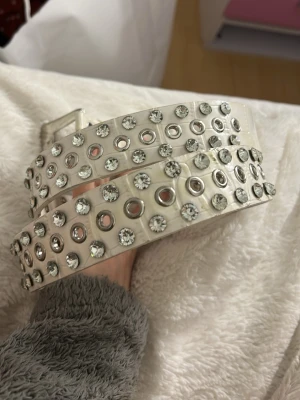 Vitt bälte med nitar och strassdetaljer - Säljer ett vitt bälte med dubbla rader av silverfärgade nitar och glittrande strassdetaljer. Spännet är dekorerat med små stenar för extra bling. Perfekt accessoar för att lyfta vilken outfit som helst.