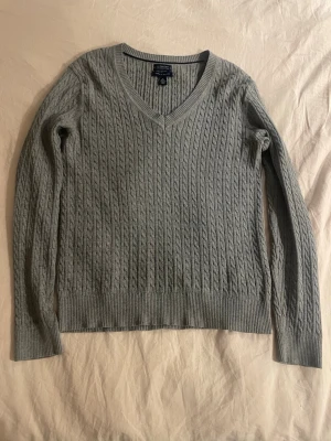 Grå kabelstickad tröja Tommy Hilfiger - Snygg grå kabelstickad tröja från Tommy Hilfiger med V-ringning och långa ärmar. Tröjan har ribbade muddar vid ärmslut och nederkant samt diskret logga på ärmen. Perfekt för lager på lager och enkel att matcha.