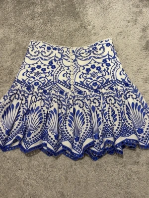 Vit och blå kjol från Zara - Supersnygg vit kjol från Zara med blått, detaljerat mönster och volangkant. Kjolen är mid waist och har knappar framtill och ett unikt, nästan boho-inspirerat print. 