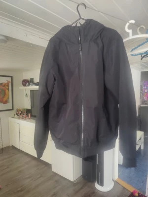 Svart vindjacka från Etirel med huva - Svart vindjacka från Etirel i storlek S. Jackan har dragkedja framtill, huva med dragsko och ribbade muddar vid ärmslut och nederkant. Perfekt för blåsiga dagar och enkel att matcha med olika outfits.