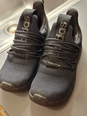 Svarta Adidas sneakers med mesh - Snygga svarta sneakers från Adidas med ovandel i meshmaterial och tydlig Adidas-logga på plösen. Skorna har låga skaft och klassisk snörning. Perfekta för en sportig och avslappnad stil.