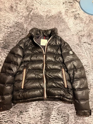 Svart dunjacka från Moncler - Snygg svart dunjacka från Moncler med glansig finish och klassisk logga på ärmen. Jackan har två fickor med dragkedja och detaljer i rött, vitt och blått längs dragkedjorna. Perfekt för kalla vinterdagar och riktigt trendig look.
