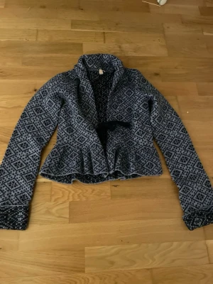 Mönstrad blå kofta med volang - Snygg blå kofta med svart och vitt geometriskt mönster. Koftan har volang nedtill, långa ärmar och en liten knytning i midjan. Perfekt för dig som gillar unika detaljer och vill ha något extra till din outfit.Använd några få gånger och i väldigt bra skick.Ordinarie pris 499kr