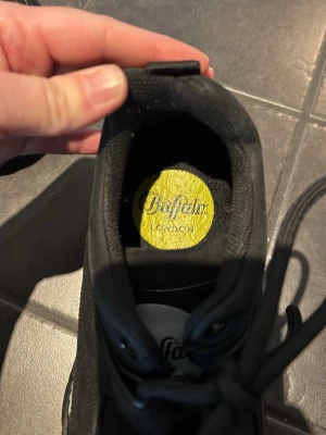 Buffalo London svarta chunky sneakers - Buffalo London sneakers med chunky sula och klassisk svart färg. Skorna har rund tå, snörning och logodetaljer på plösen och innersulan. Materialet är syntet och textil, perfekt för dig som gillar en edgy och trendig stil.