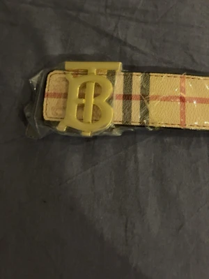 Rutig Burberry-inspirerat bälte - Snyggt bälte med klassiskt rutigt mönster i beige, svart och rött samt ett stort guldfärgat spänne med bokstäver. Perfekt accessoar för att lyfta din outfit och ge en exklusiv känsla. Helt ny aldrig använd, plasten är kvar. Skickas samma dag eller dagen efter köp, MVH.