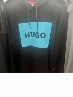 Svart hoodie från HUGO med blå logga - Snygg svart hoodie från HUGO med stor blå logga på bröstet. Klassisk huva med dragsnören och en stor magficka framtill. Perfekt för en avslappnad och trendig stil. Tillverkad i mjukt material för extra komfort.